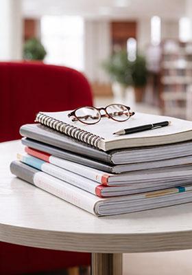 Stack of academic notebooks and research documents with glasses and a pen on a table in a university library.